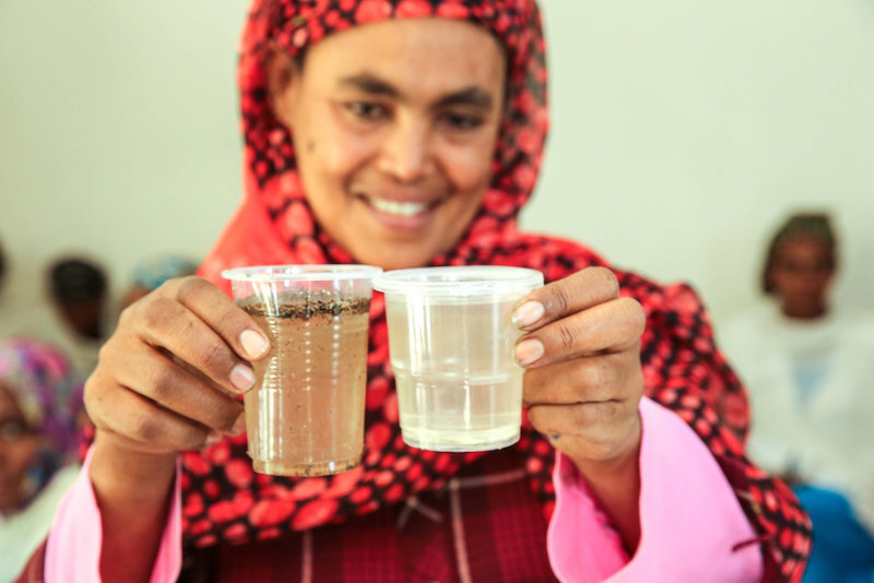 a woman holds up a dirty cup of water next to a clean cup of water