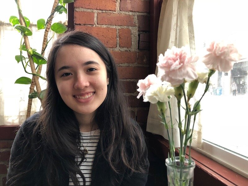 girl sits smiling against a brick background near a vase of flowers