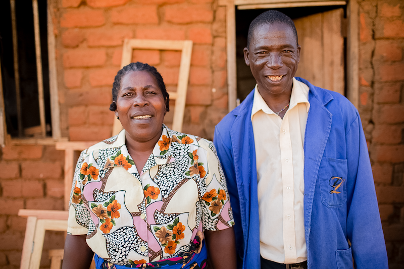 a man wearing a blue workman’s coat stands smiling side-by-side with his wife