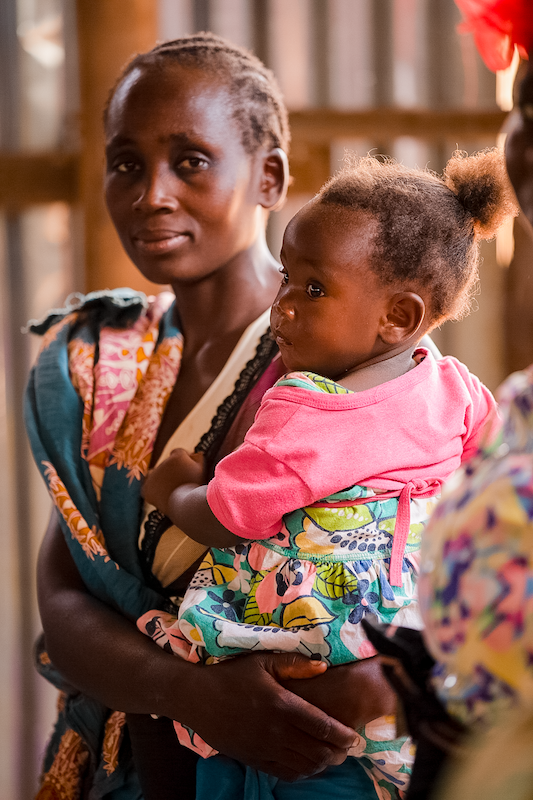 woman stands in colorful clothing, holding a young child